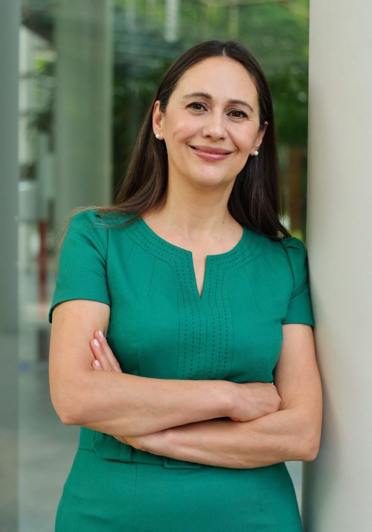 Frau mit langen, dunklen Haaren und grünem Oberteil, lächelt mit verschränkten Armen.
